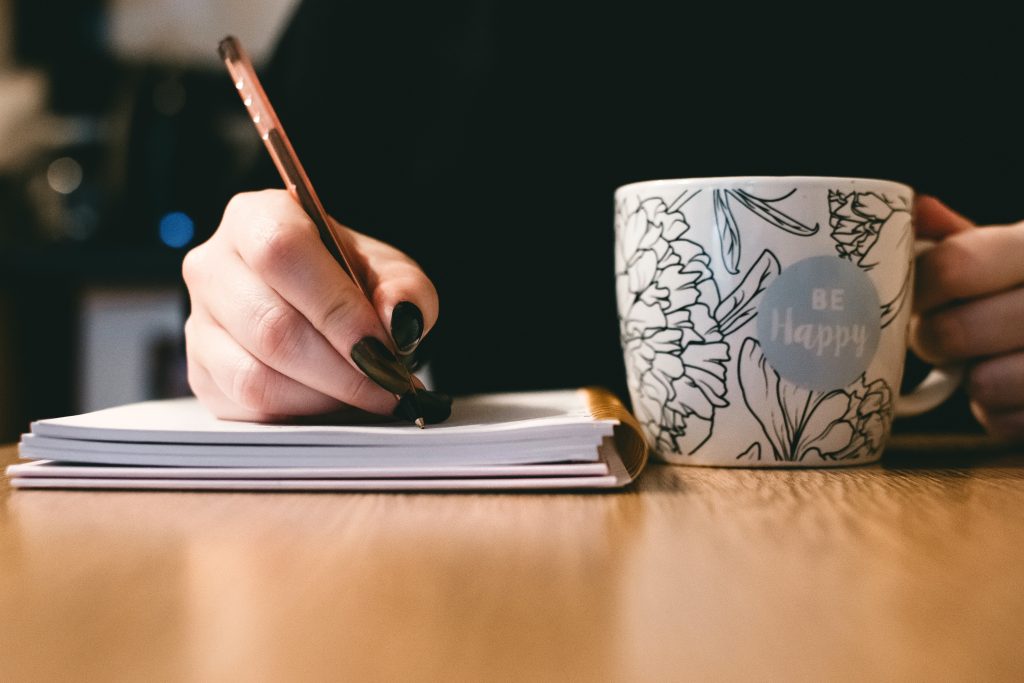 Schreibende Frau mit Kaffeetasse mit Schriftzug "Be happy". Foto: Pexels, Lisa Fotios